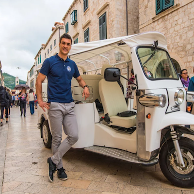 Voici les balises alt pour les 3 images : 1. TukTuk-Dubrovnik-1.png (chauffeur en polo bleu devant un tuktuk blanc à Dubrovnik) Chauffeur de tuktuk électrique blanc dans les ruelles de Dubrovnik